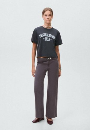 Graues, kurzes T-Shirt mit dem Aufdruck "SISTERHOOD USA 2005"; kombiniert mit weiten, braunen Hosen, die zwei Knöpfe und eine glatte Oberfläche haben.