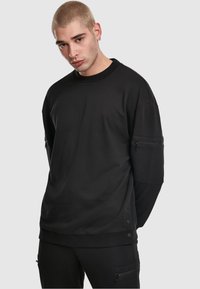 Felpa nera dal taglio ampio, con scollo rotondo, tasche laterali con cerniera e dettagli con bottoni automatici sull'orlo. Texture liscia.