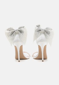 Chaussures à talons hauts blanches avec une finition satinée, ornées de grands nœuds en sequins argentés à l'arrière et d'accents en perles sur les brides.