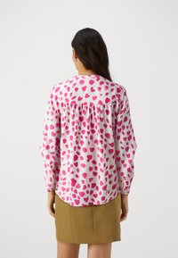 Blusa a maniche lunghe in rosa e bianco, con motivo a cuori, dettaglio di schiena arricciata e scollatura rotonda. Il tessuto sembra leggero e liscio.