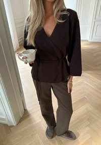 Dunkelbraunes Wickeltop mit weiten Ärmeln, das an der Taille gebunden ist. Kombiniert mit braunen weiten Hosen und grauen Slippern. Hält eine weiße Keramiktasse.