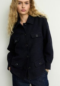 Navy textured jacket with a collar, featuring two chest pockets, black buttons, and a relaxed fit, paired with dark denim jeans.