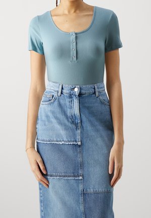 Frau trägt ein tailliertes, geripptes hellblaues Kurzarmshirt mit Knöpfen, kombiniert mit einem Patchwork-Denimrock.