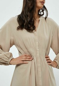 Abito beige con scollatura a V e vita arricciata. Maniche lunghe a sbuffo con polsini elasticizzati. Tessuto dalla texture liscia con un leggero lucido.