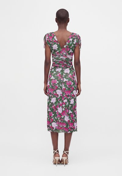 Femme debout de dos, portant une robe verte longueur genou avec un imprimé floral rose et blanc et des sandales beiges à talons hauts.