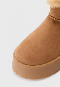 Bottine en suede marron avec un bout rond, présentant une semelle intermédiaire texturée et un accent tressé près du bord supérieur. Doublure intérieure douce.