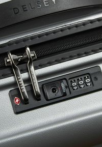 Delsey suitcase zipper area features dual silver metal pulls, a combination lock, and a textured silver surface with security system detail.