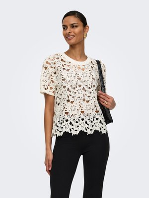 Femme souriante, portant un haut blanc en dentelle à fleurs à manches courtes, un pantalon noir, des boucles d'oreilles en or, une bague, et portant un sac à bandoulière noir.