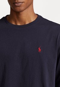 Marineblaue Baumwoll-T-Shirt mit rundem Ausschnitt und kleinem roten gesticktem Logo auf der linken Brust. Glatte Textur, normale Passform.