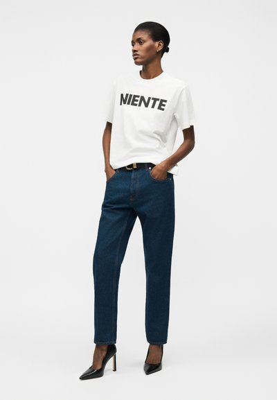 Γυναίκα που φοράει λευκό t-shirt με τη λέξη "NIENTE", σκούρα μπλε τζιν με μαύρη ζώνη και μαύρες ψηλοτάκουνες γόβες, στέκεται με τα χέρια στις τσέπες.