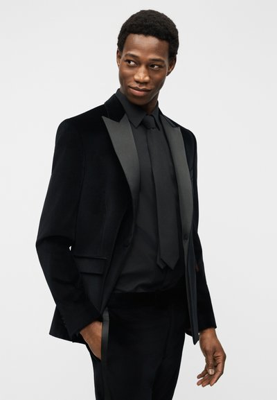 Άνδρας με μαύρο βελούδινο σμόκιν με σατέν πέτα, μαύρο πουκάμισο και γραβάτα, στέκεται με το ένα χέρι στην τσέπη, κοιτάζοντας πλάγια με ελαφρύ χαμόγελο.