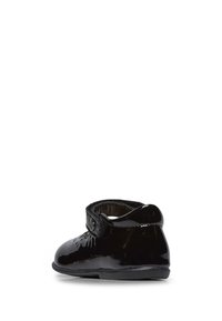 Zapato de charol negro con punta redonda, suela plana y una correa de velcro. Presenta costuras decorativas en la zona de la punta y un forro suave.