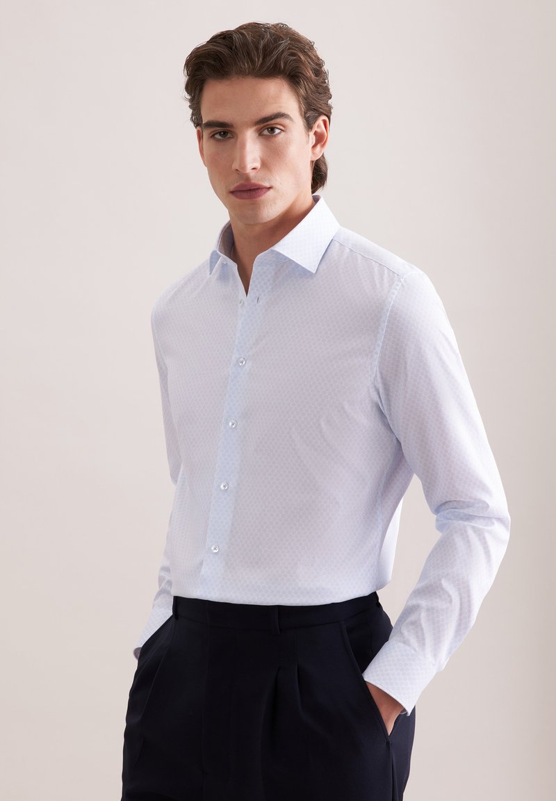 Jeune homme portant une chemise habillée à motifs bleu clair, rentrée dans un pantalon noir plissé, les mains dans les poches, fond neutre.