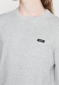 Maglione a coste grigio con scollo a girocollo, caratterizzato da un'etichetta logo nera di Calvin Klein sul petto. Morbido al tatto.