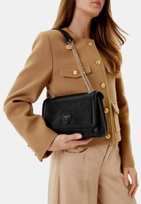 Borsa nera con texture, dotata di un logo triangolare e una tracolla a catena, abbinata a un giubbotto color cammello con bottoni dorati.