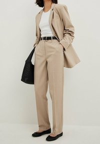 Beige skræddersyet blazer over en hvid top, parret med løse beige bukser. Tilbehør inkluderer et sort bælte og en vævet sort håndtaske.