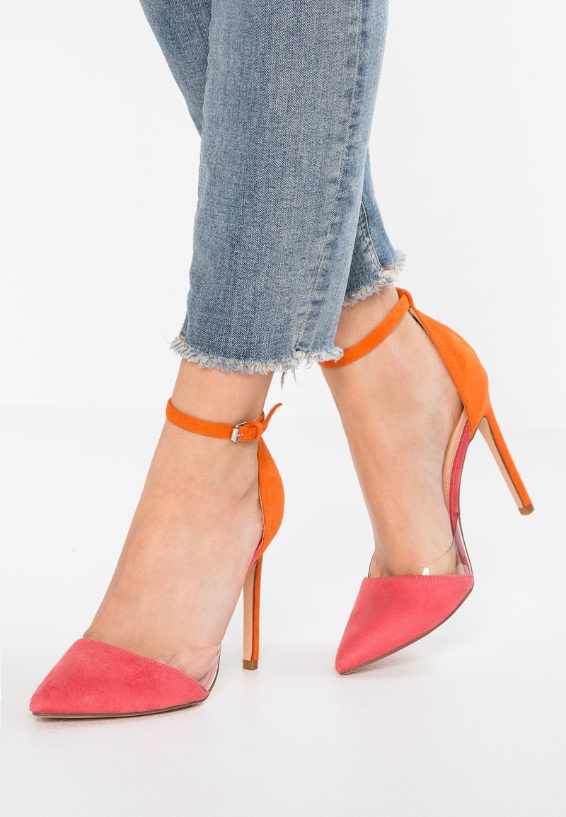 Pieds portant des escarpins à bout pointu rose et orange avec des brides à la cheville, associés à un jean bleu à ourlet effiloché.