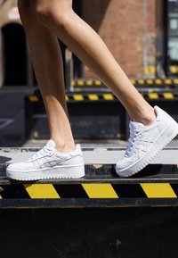 White leather sneakers with a textured surface, round toe, and a thick white sole. Feature three diagonal stripes on the side.
