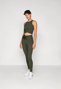 Donna in piedi che indossa un crop top senza maniche verde scuro, leggings a vita alta abbinati, un cappellino nero e sneakers bianche su uno sfondo semplice.