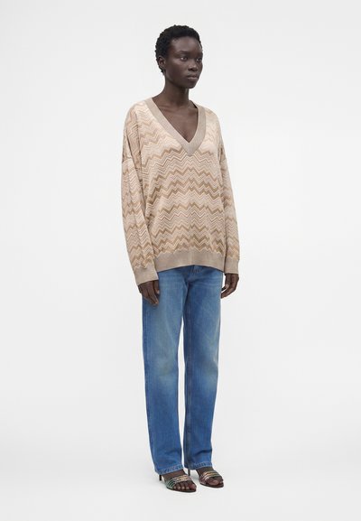 Personne portant un pull beige à motif zigzag en V, un jean bleu droit et des sandales à talons noires avec des brides colorées.
