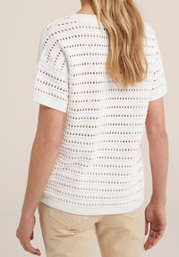 Vit stickad tröja med korta ärmar, med ett horisontellt randigt mönster och ett öppet arbetat mönster, avslutad med en ribbad fåll vid midjan.