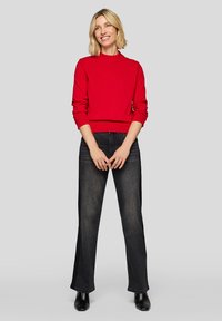 Roter Pullover mit hohem Kragen und langen Ärmeln, kombiniert mit dunkel waschgebleichten Jeans mit weitem Bein und schwarzen Stiefeletten vor einem grauen Hintergrund.