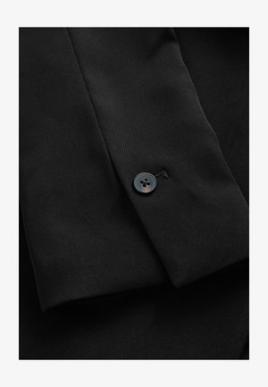 Juodas audinys su raukšlėta tekstūra, ant rankovės matosi vienas mygtukas. Tvarkingas, pritaikytas stilius.