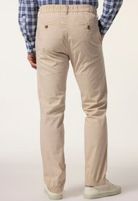 Beige elastiska byxor med avslappnad passform. Har två bakfickor med knappstängning och ribbade muddar vid ankeln.