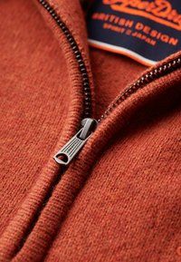 Indumento zip-up di colore rosso-arancione con texture, dotato di una zip in metallo e di un'etichetta in tessuto con la scritta "British Design. Spirit & Japan."