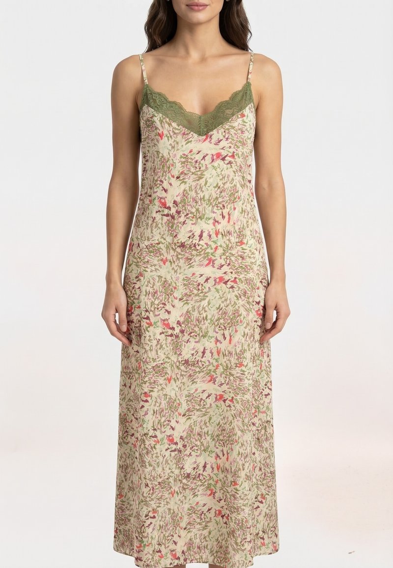 Woman wearing sleeveless beige maxi dress with green lace trim and pink, green, and purple abstract floral pattern, standing with arms relaxed.