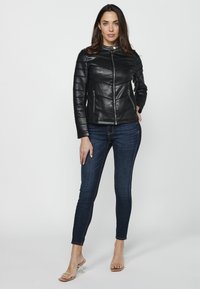 Veste en cuir noir avec des manches matelassées, un col haut et des zips à l'avant, associée à un jean skinny bleu foncé et des sandales à talons transparents.