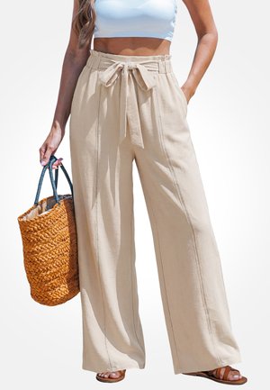 Frau trägt beige, hochtaillierte, weitgeschnittene Hose mit Bindegürtel, weißes Crop-Top, hält eine geflochtene Strohtasche und trägt braune Sandalen.