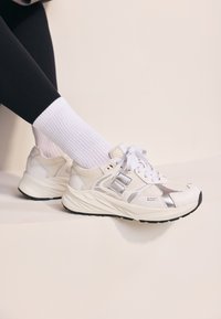 Scarpe da ginnastica bianche e argentate con pannelli in rete, sovrapposizioni testurizzate e una suola ammortizzata; indossate con calze bianche a costine e leggings neri.
