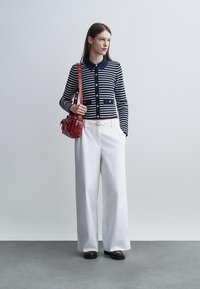 Femme portant un cardigan boutonné rayé bleu marine et blanc, pantalon blanc large, chaussures noires, portant un sac à bandoulière en cuir rouge.