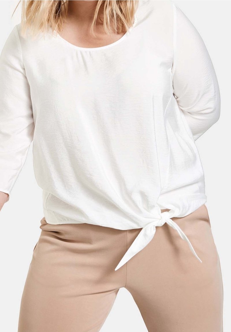 Haut blanc à manches longues noué à la taille, porté avec un pantalon beige, montrant le haut du corps du cou aux hanches.