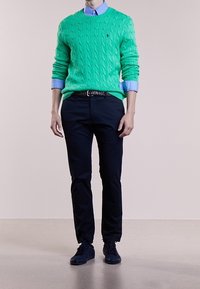 Grön stickad tröja med kabelmönster, ljusblå skjorta med krage under, matchad med mörk marinblå byxor och svarta sneakers.