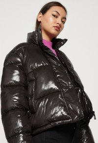 Chaqueta acolchada negra y brillante con un alto cuello, que cuenta con un grueso acolchado, cierre de cremallera frontal y botones a presión, combinada con un suéter púrpura debajo.
