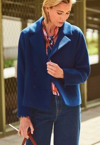 Blauwe wolmixjas met een brede kraag en dubbele knoopsluiting, gecombineerd met een kleurrijke gebloemde blouse en denim broek.