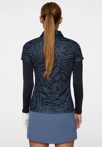 Krótki rękaw, granatowa koszulka golfowa z abstrakcyjnym wzorem topograficznym i stójką, w połączeniu z jasnoniebieską spódnicą golfową. Widoczna biała rękawiczka.