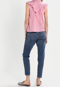 Top rosa senza maniche con dettagli a volant sul retro, abbinato a jeans di denim blu a vita alta e scarpe slip-on grigie con decorazioni.