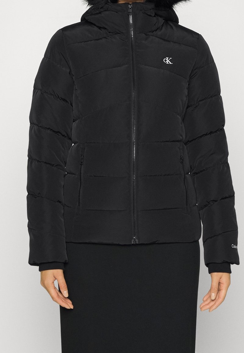 Veste d'hiver noire matelassée avec capuche, fermeture éclair avant, poches latérales zippées et logo blanc "cK" sur la poitrine.