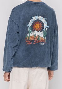 Blue oversized sweatshirt made of cotton, featuring a graphic design of a sun, mountains, and cacti with the text "Feel the Sunset."