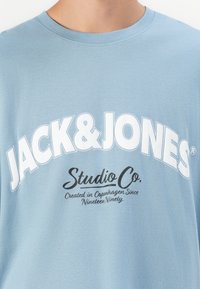 Lyseblå T-shirt med stor hvid "JACK & JONES" tekst og mindre sort "Studio Co. Skabt i København Siden nitten nitte."