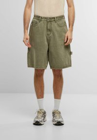 Shorts di denim verde oliva con una vestibilità comoda, cuciture visibili e tasche frontali. Indossati con una maglietta in rete color crema e sneakers.