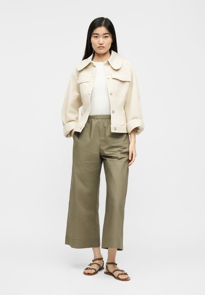 Ženska z dolgimi ravnimi črnimi lasmi, oblečena v kremno kratko jakno, belo majico, olivno široke hlače in rjave ravne sandale z naramnicami.