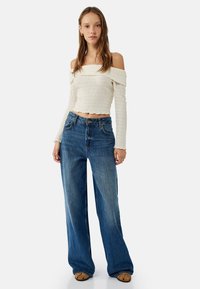 Valkoinen olkaimeton lyhyt yläosa, jossa on teksturoitu kangas, yhdistettynä leveälahkeisiin sinisiin farkkuihin. Farkuissa on klassinen viiden taskun muotoilu.