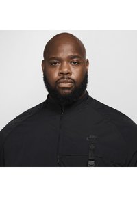 Zwarte lichte jas met een hoge kraag, ritsluiting en voorkantzak. Heeft een discreet Nike-logo op de borst.