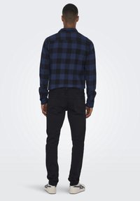 Camicia a scacchi blu e nera con maniche lunghe, taglio dritto e jeans neri. Modello che indossa sneaker bianche. Vista da dietro.