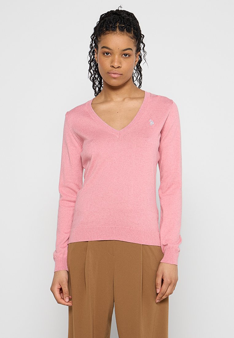Maglione rosa a V con maniche lunghe, texture morbida, polsini e orlo a coste, con un piccolo logo sul lato sinistro del petto. Pantaloni marroni indossati in basso.