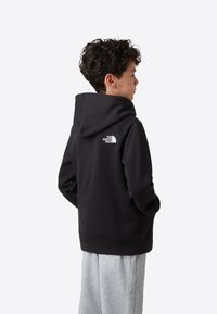 Črna kapucar z teksturirano površino in belim logotipom na hrbtu. Kombinirana z svetlo sivimi trenirkami.